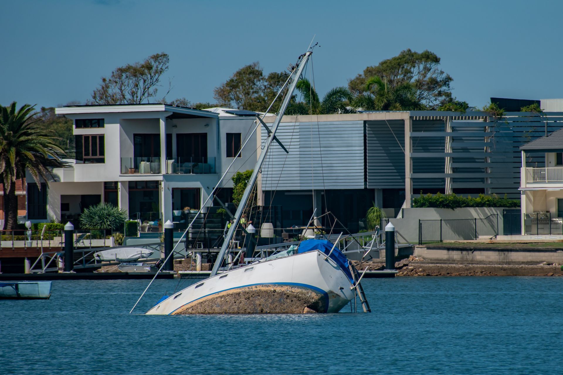 Capsized white sailboat in blue water near modern waterfront buildings, with a tilted mast and rigging visible