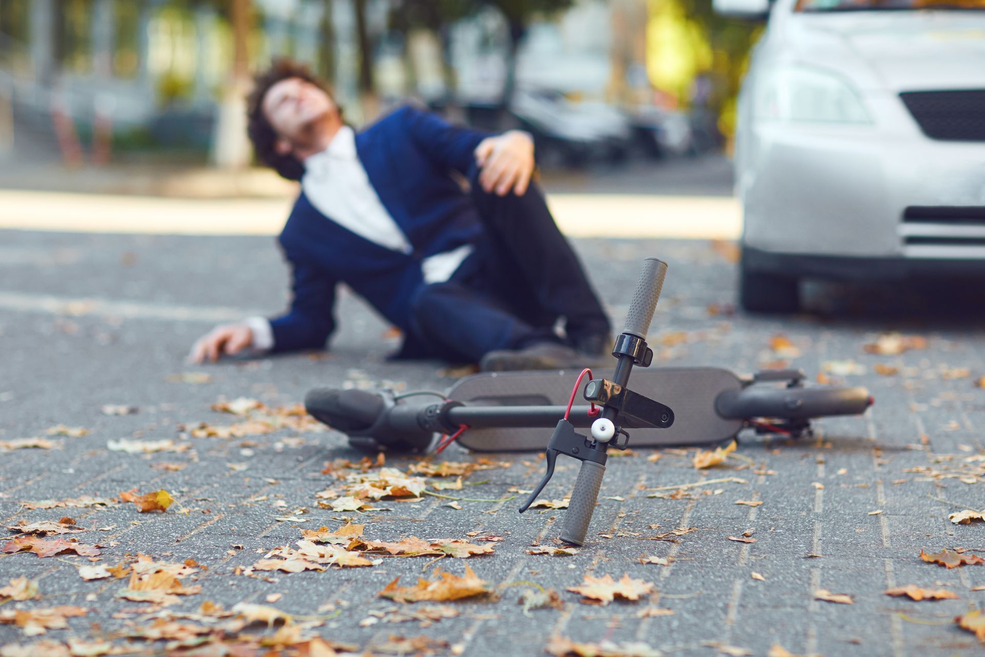 Bicycle lying on road beside a fallen cyclist and a parked car on a leaf-covered street