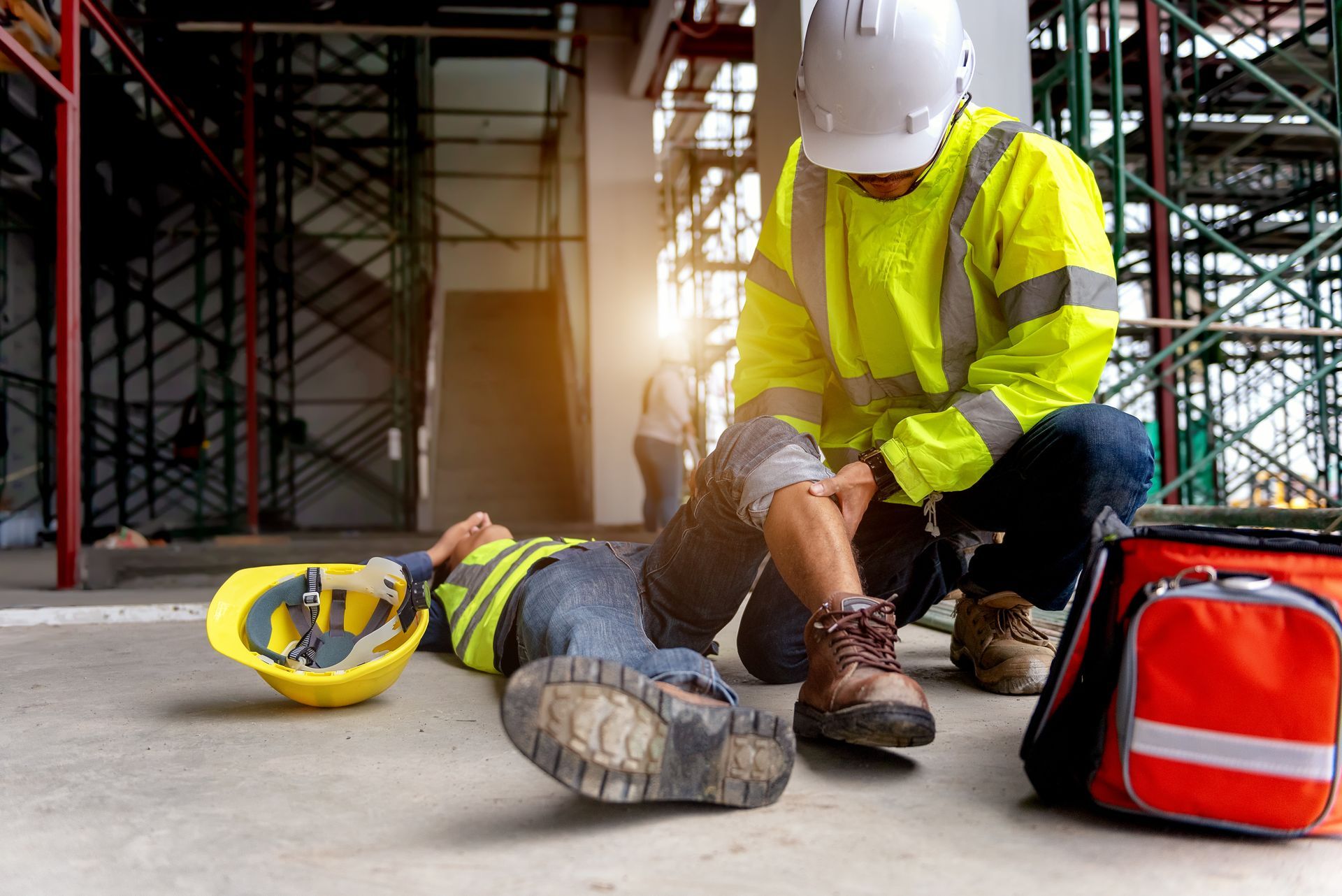 Construction worker tending to injured colleague on site, with hard hat and red first aid bag nearby