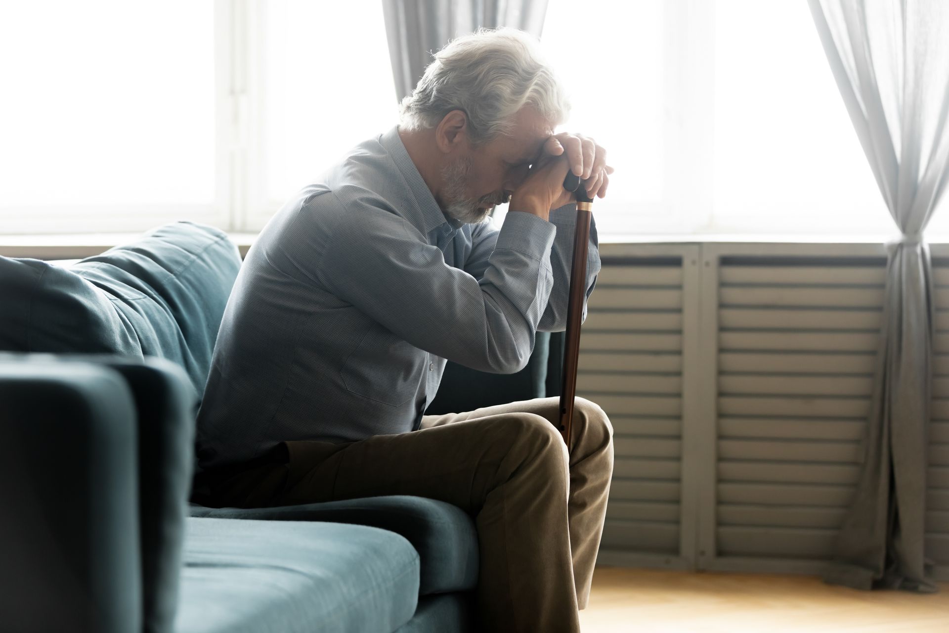 Person sitting on a couch, leaning on a cane and resting their head in their hand by a bright window.