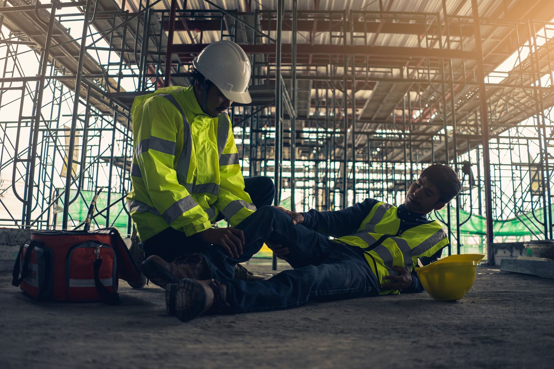 Two construction workers in high-vis gear and helmets assist an injured worker on a building site.