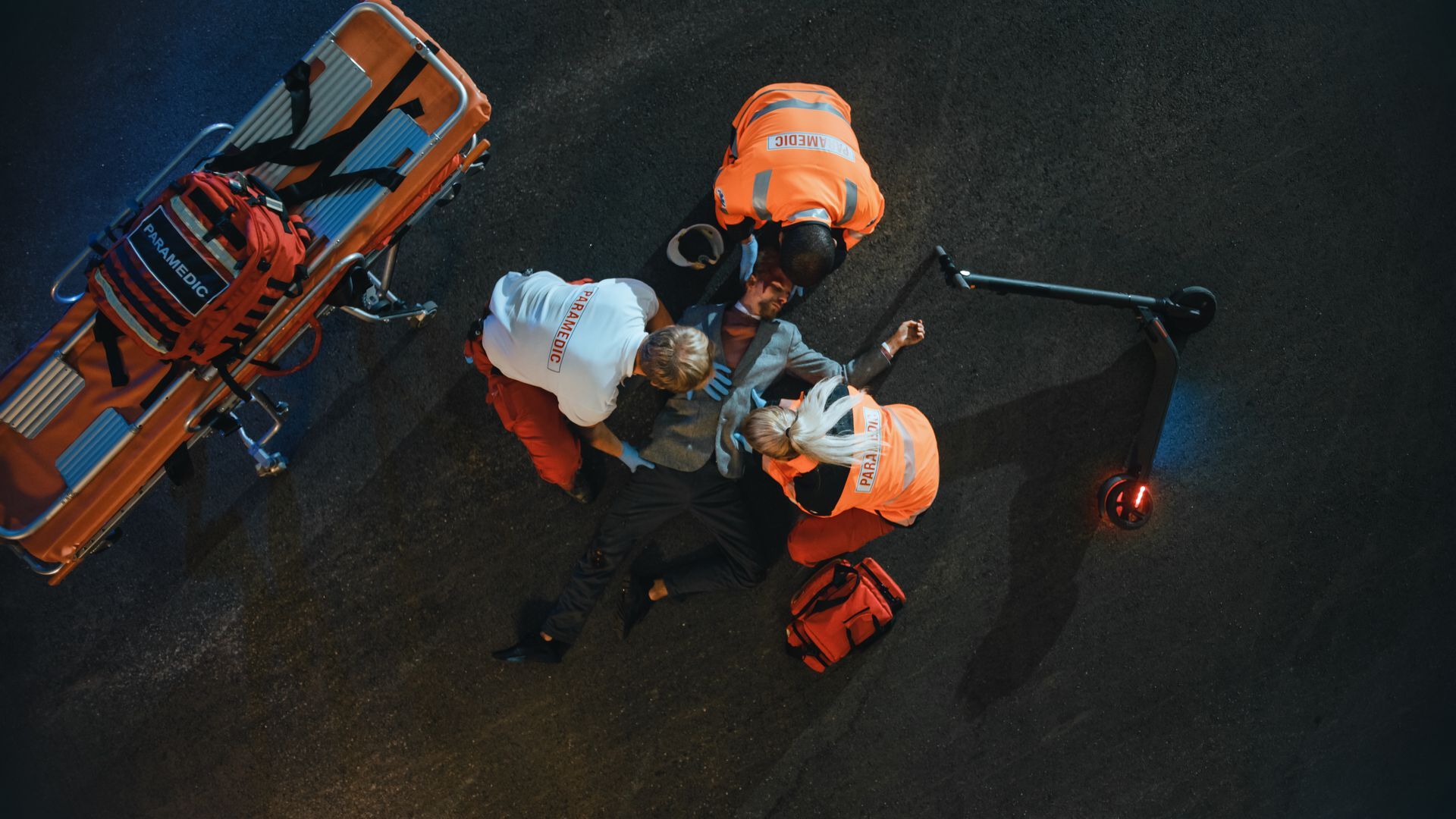 Aerial view of workers in orange safety gear gathered around a stretcher on a dark road at night