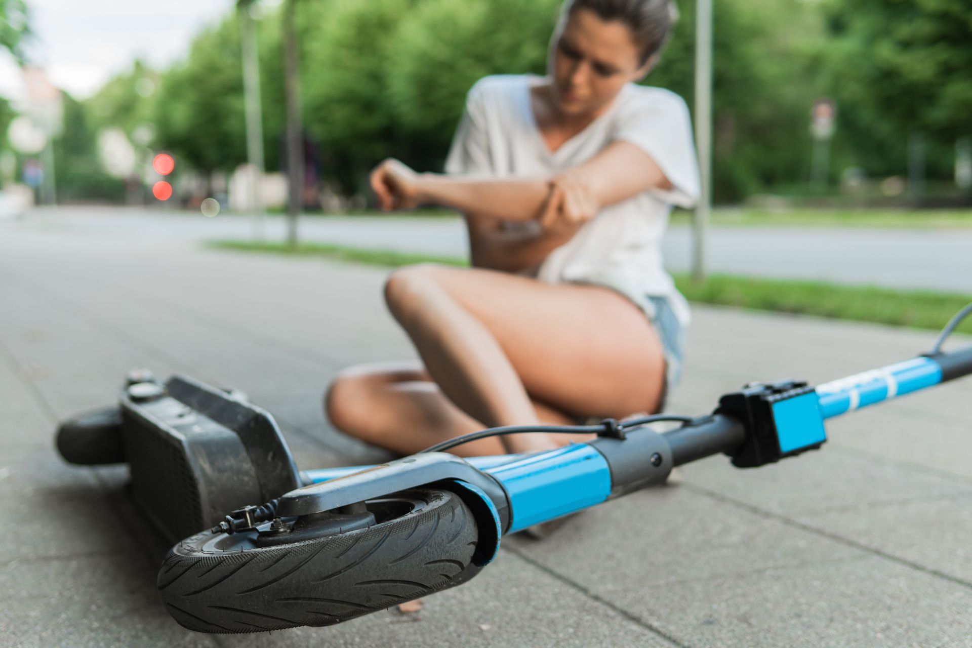 Person sitting beside a blue electric scooter on a sidewalk, holding their injured leg.