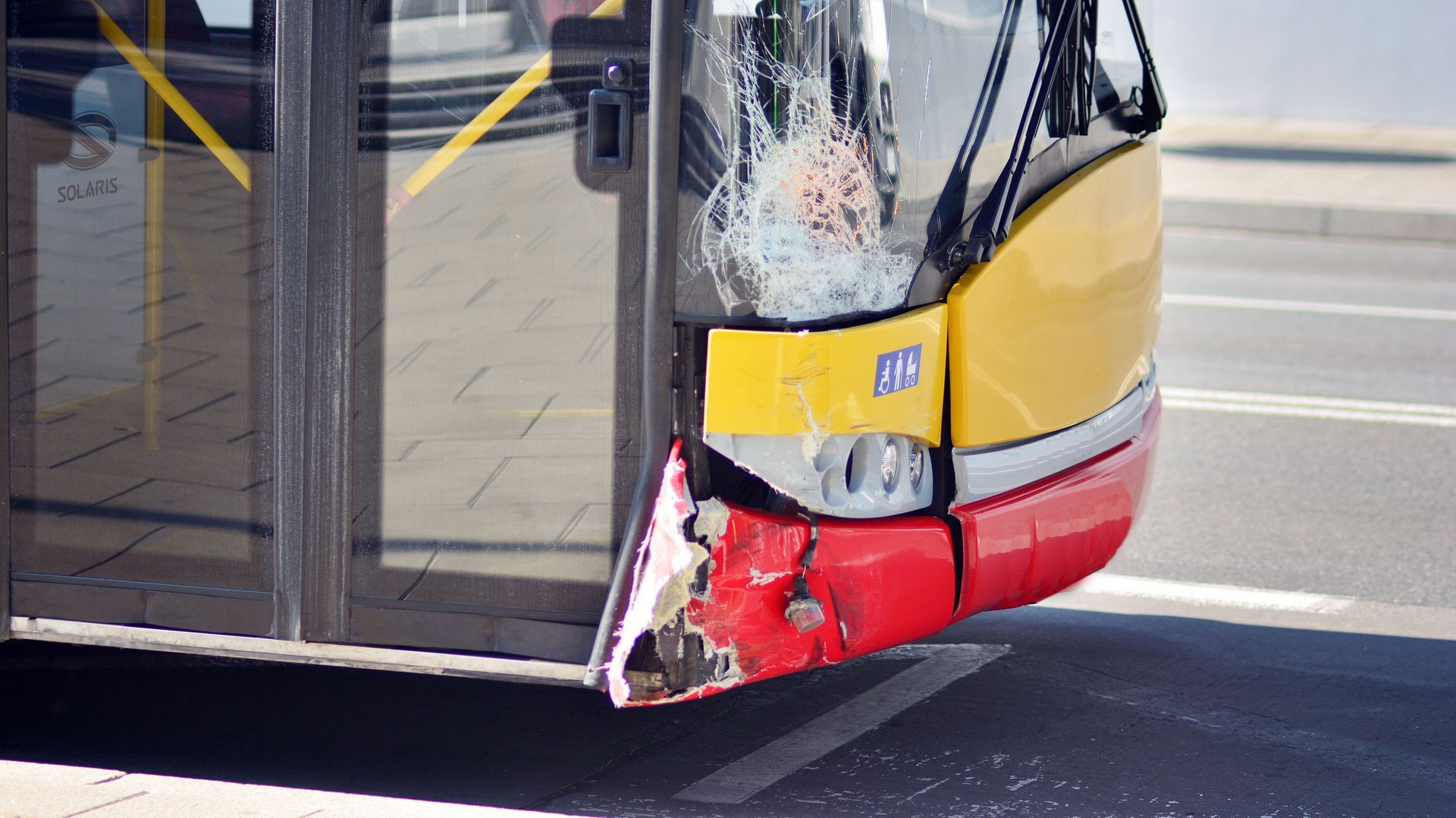 Damaged yellow-and-red bus front corner with shattered windshield at a roadside stop