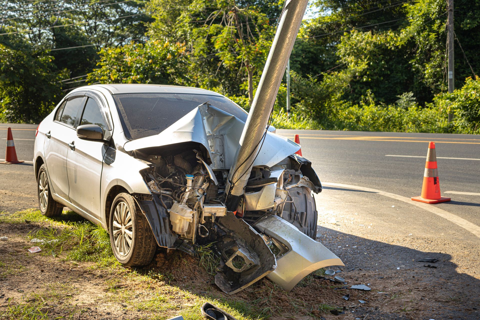 Silver car crashed into a utility pole on a roadside, front end badly damaged, with traffic cones nearby
