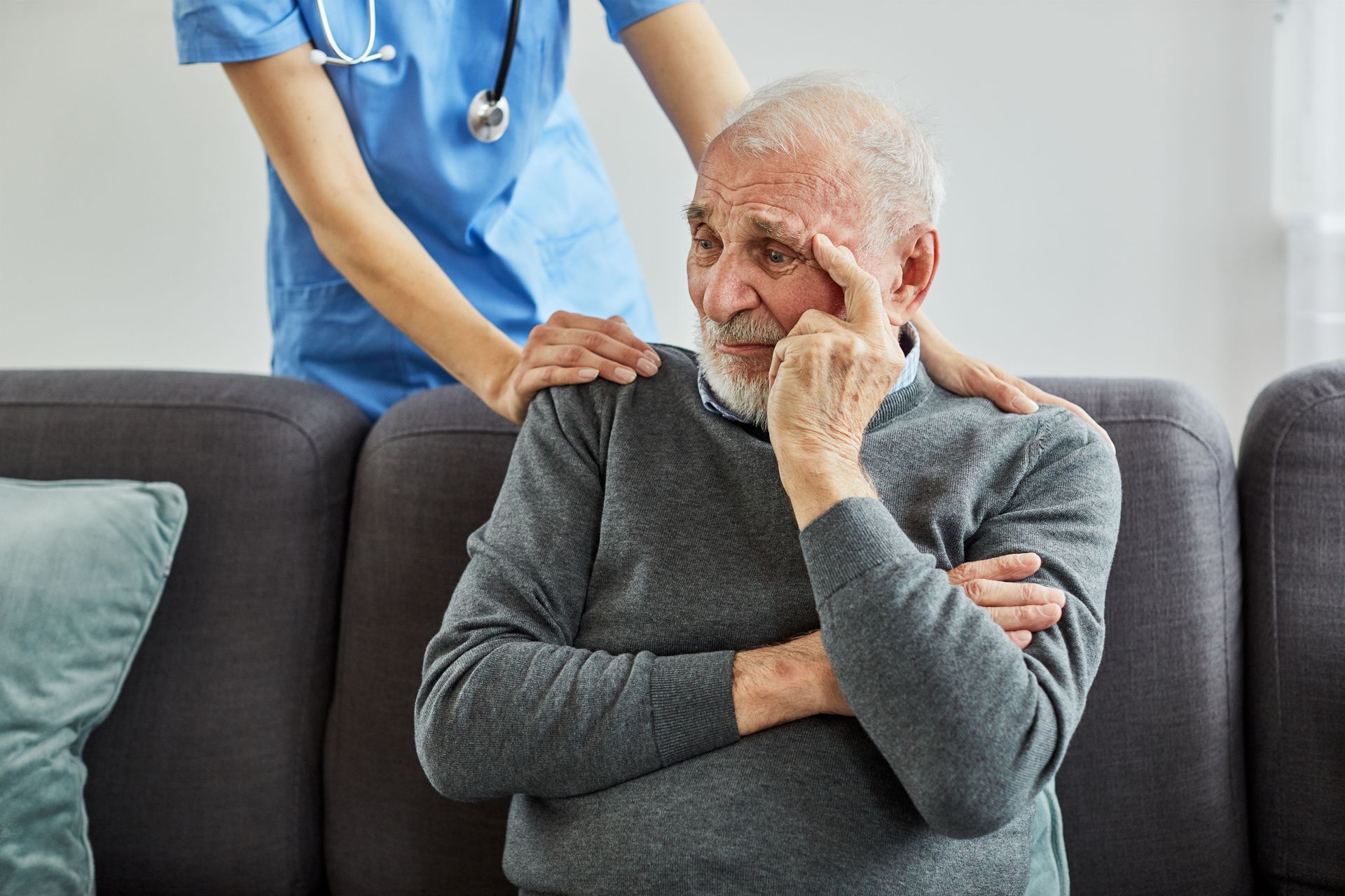 Elderly man sitting on a couch with a caregiver standing behind, hands on his shoulders in a caring gesture.