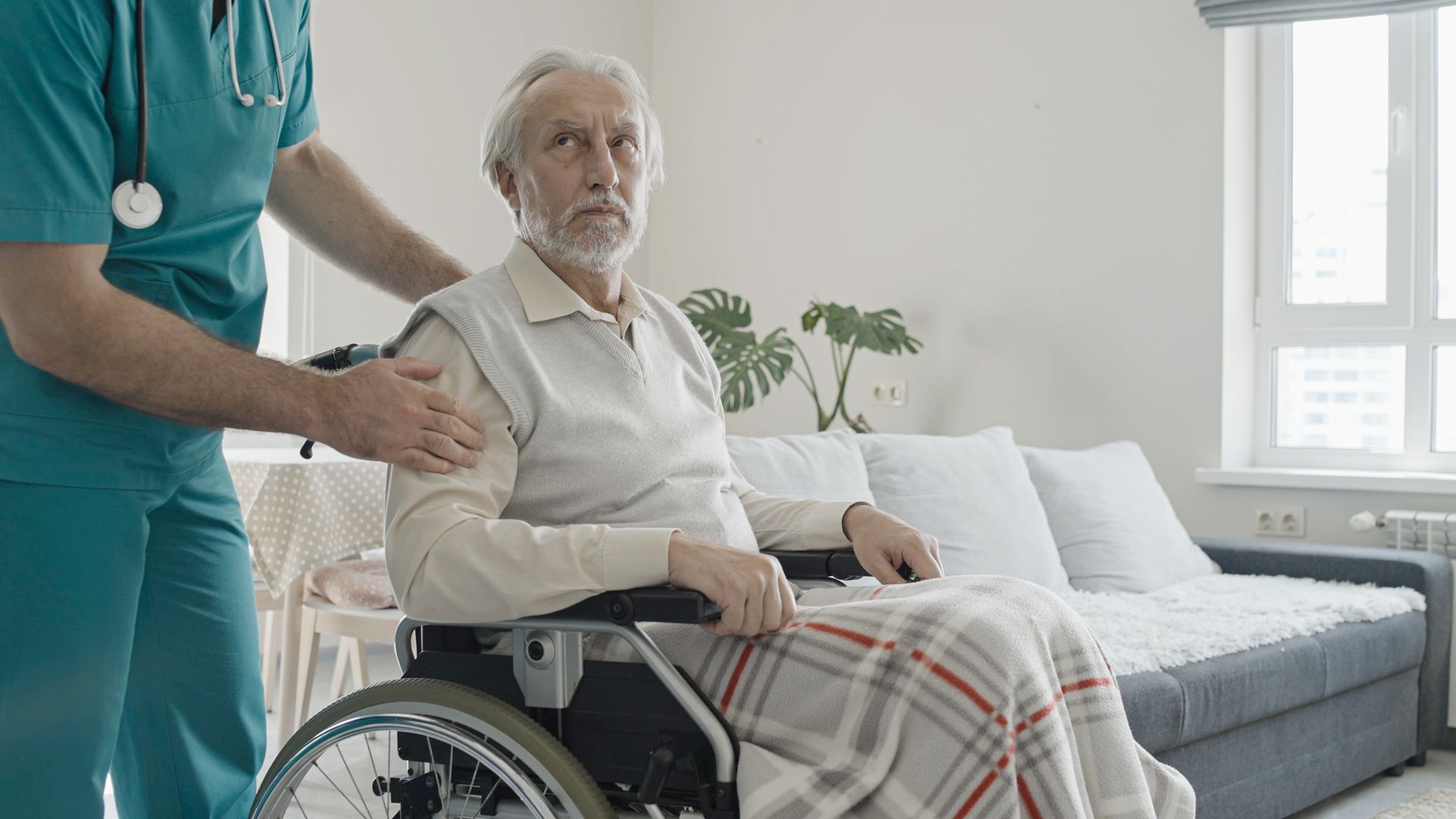 Elderly patient in a wheelchair with a caregiver in teal scrubs in a bright living room