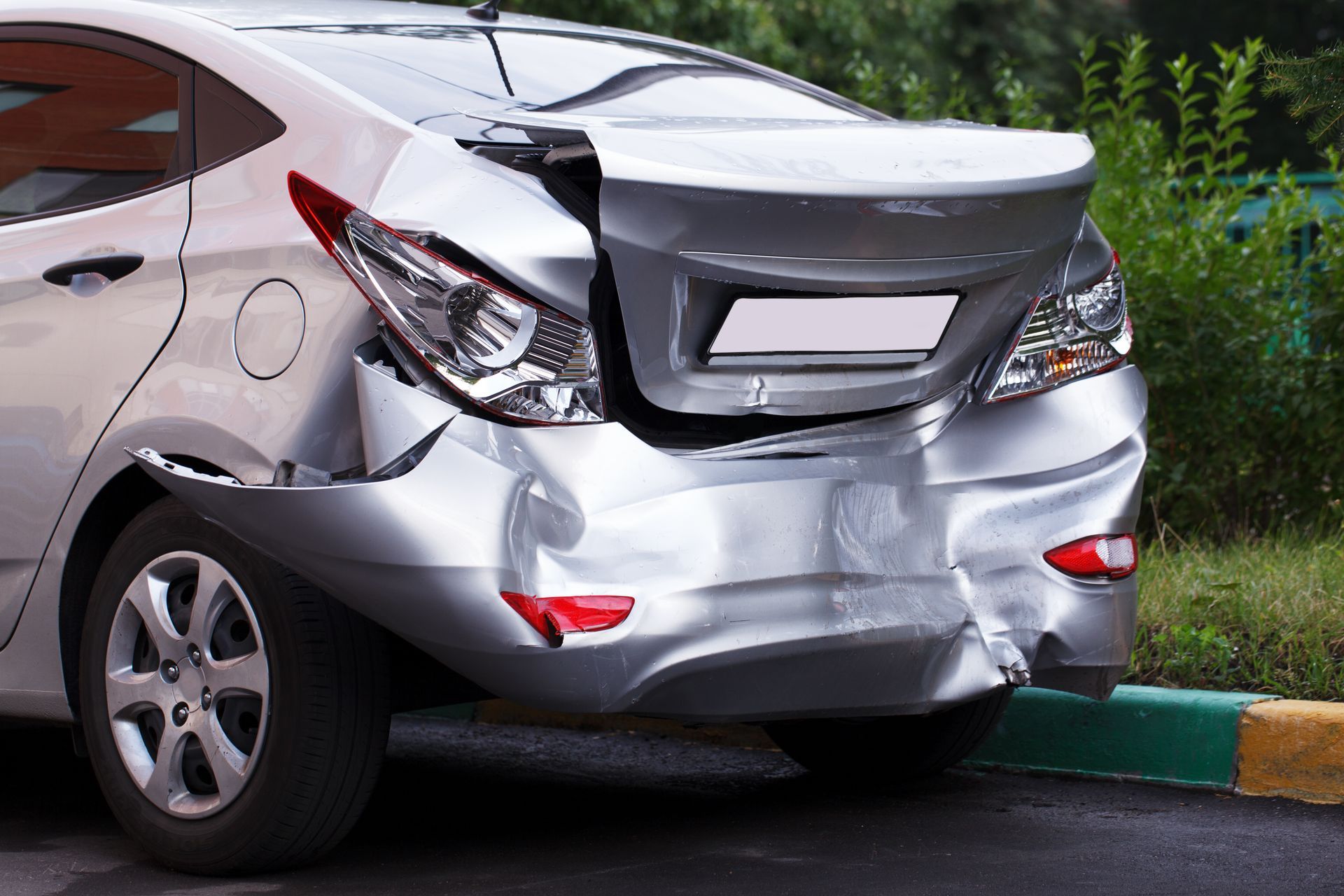Rear-end damage on a silver sedan parked by a curb, with a crumpled trunk and bumper