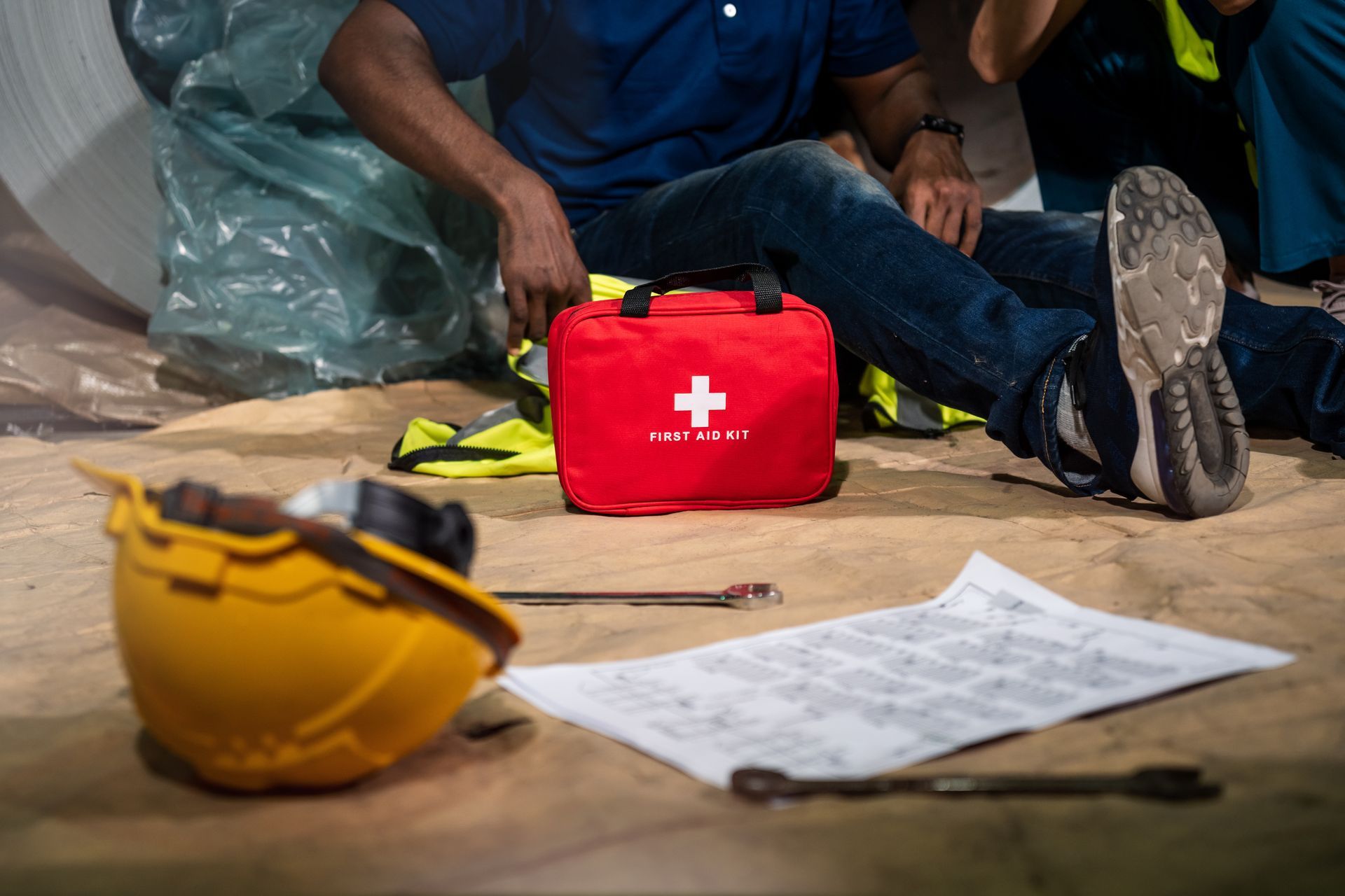 Red first aid kit on the floor beside a yellow hard hat and papers, with a seated worker in the background