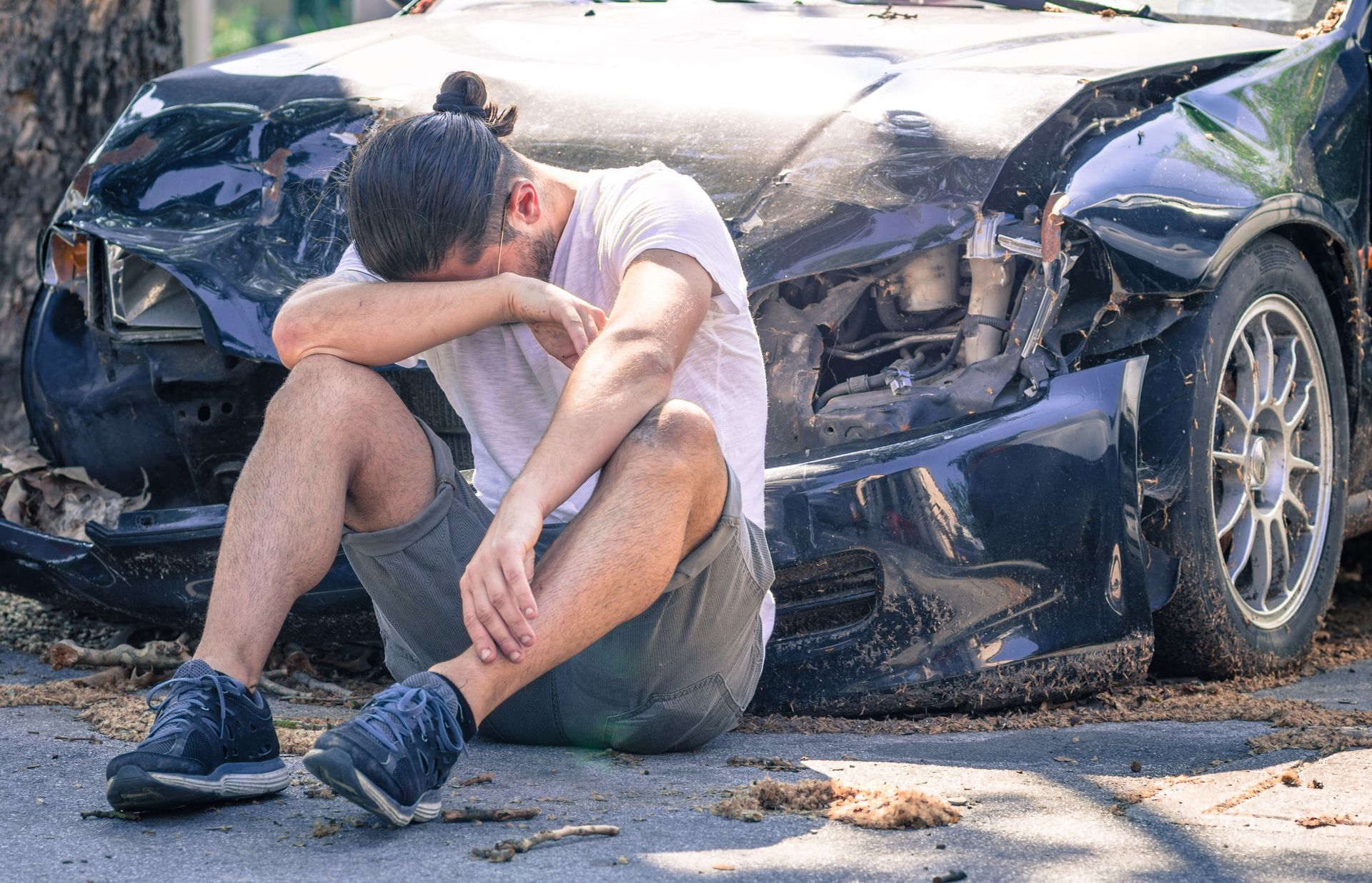 Man sitting on a sidewalk with head in hands beside a heavily damaged black car after a crash