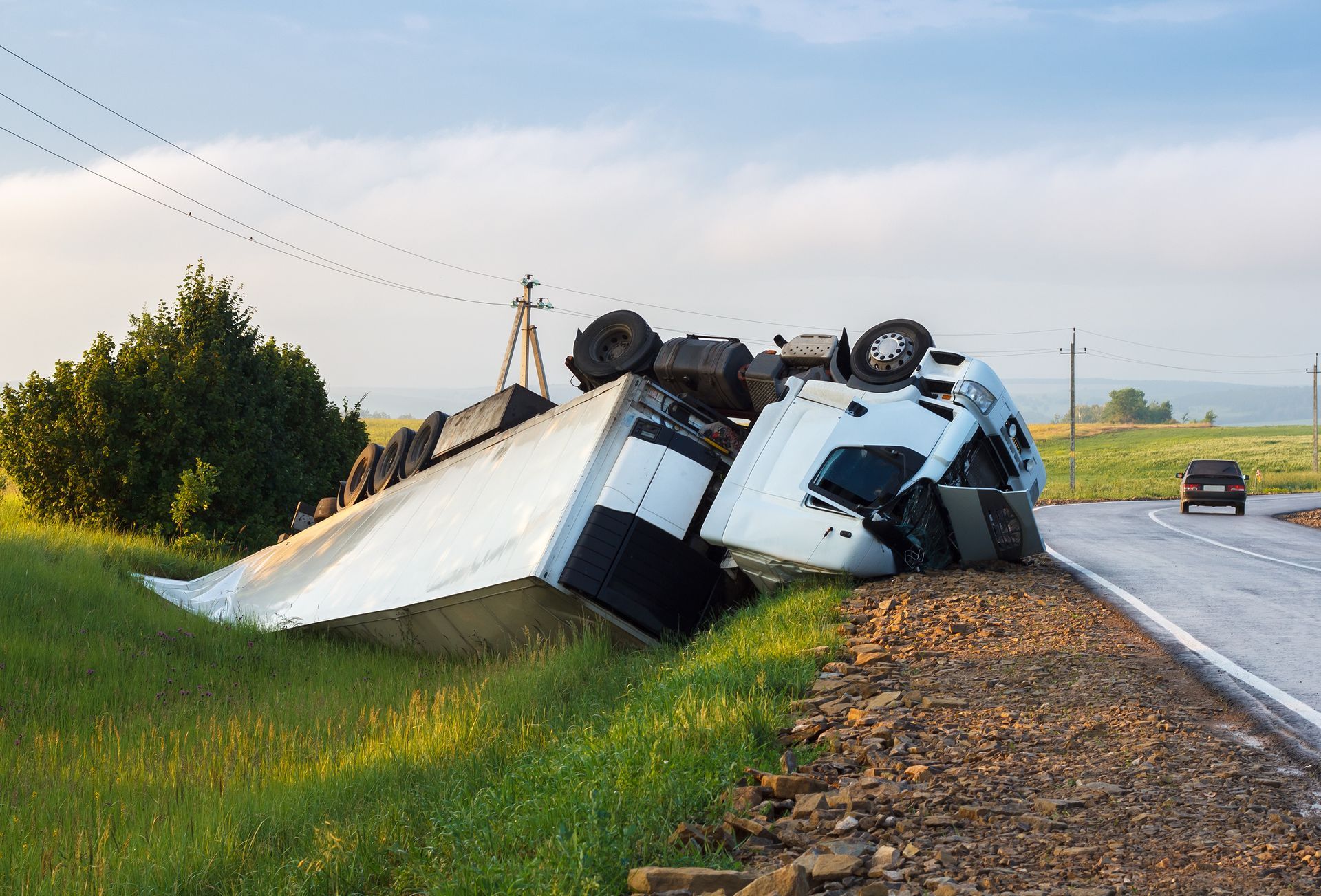Overturned white truck on its side beside a rural road at sunrise, with spilled cargo and fields nearby