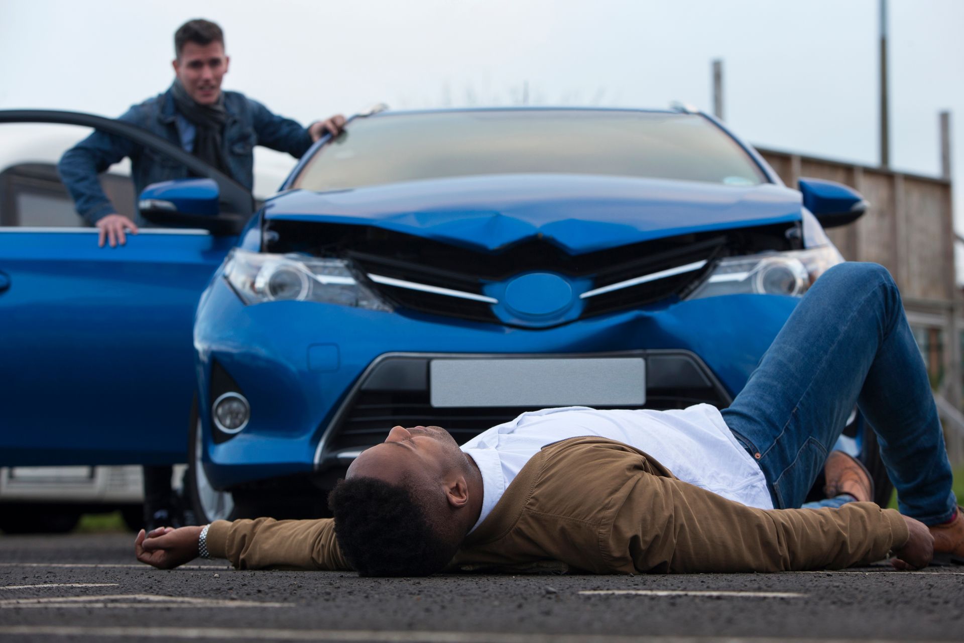 Man lying in front of a blue car while another man stands by the open door in a street scene