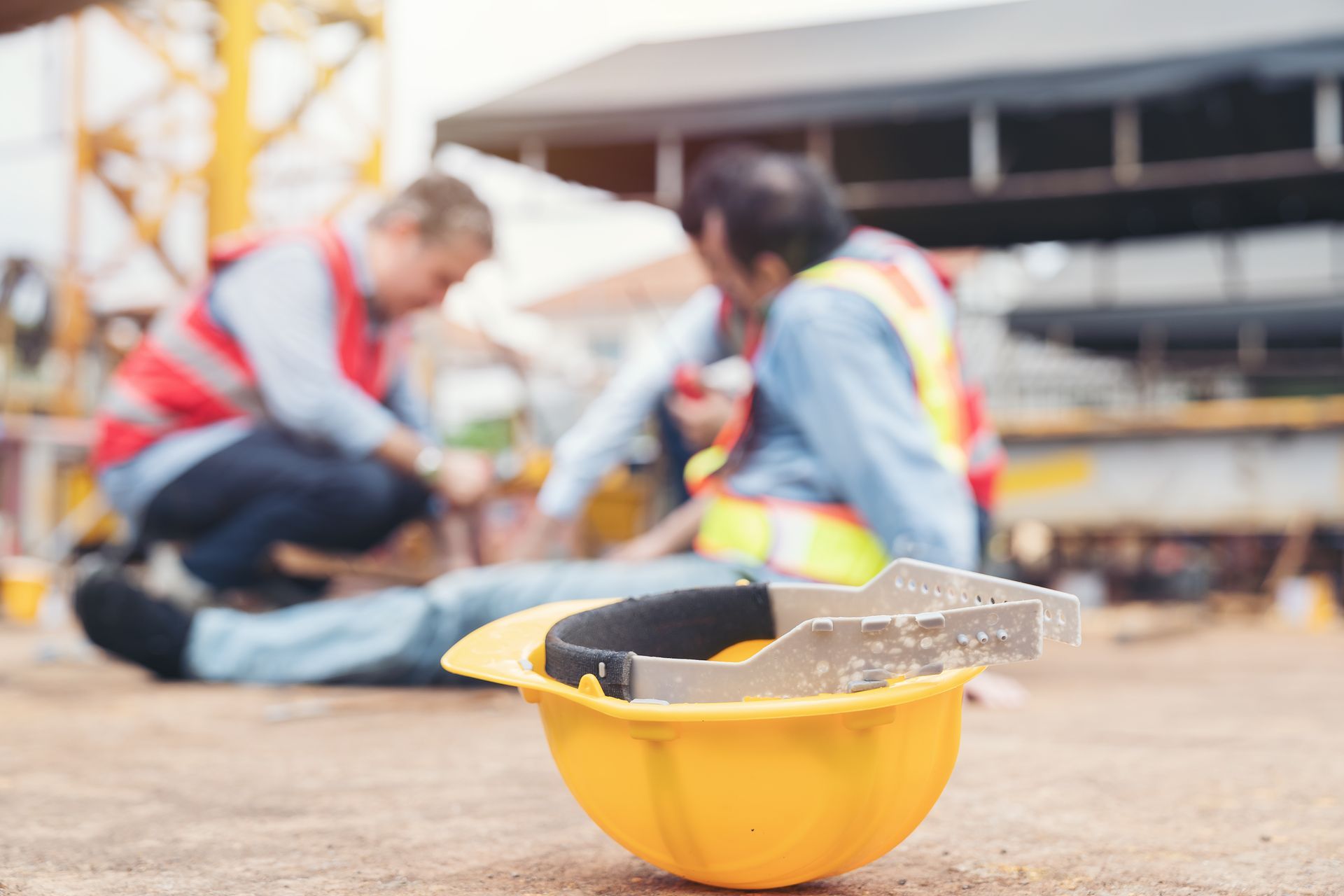 Yellow hard hat with tools in foreground; blurred construction workers kneel at a building site.