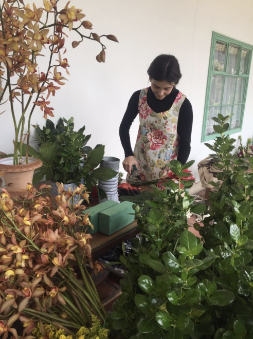 A woman is standing in a room surrounded by plants and flowers.