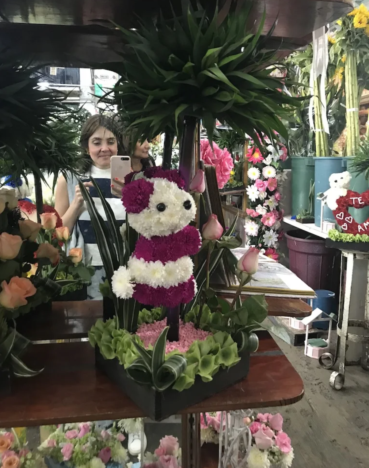 A woman is taking a picture of a teddy bear made of flowers in a flower shop.