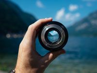 Hand holding a camera lens pointed at a blue mountain lake and sky