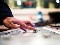 Hand adjusting audio mixer controls in a studio console, with blurred equipment in the background