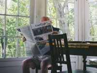 Person reading a newspaper at a table by large windows in a bright room