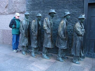 Person in blue jacket standing beside a row of bronze statues on a sidewalk outside a brick wall.