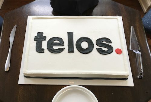 Rectangular white cake with black “telos.” lettering and a red dot on a table with plates and utensils