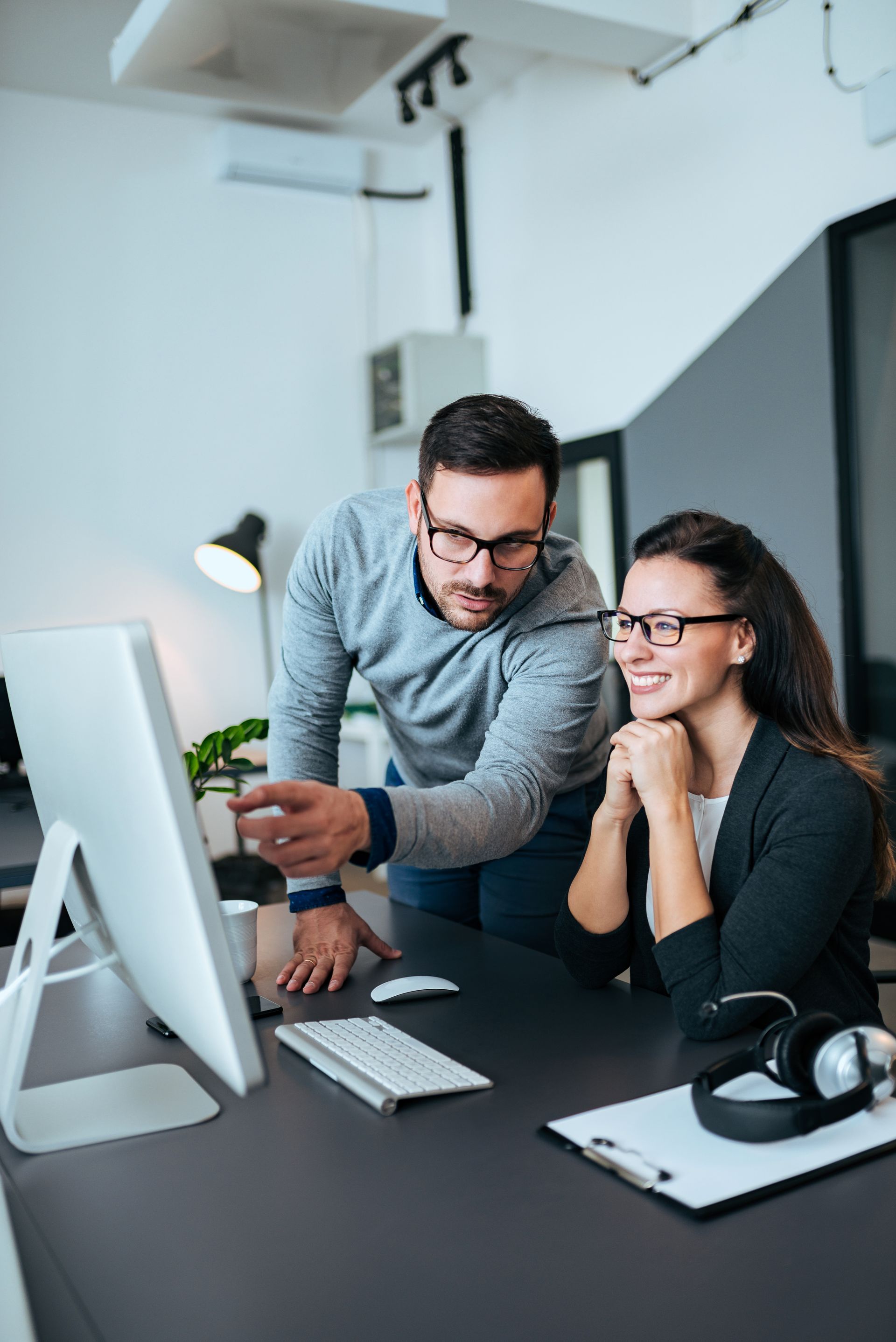 Man points at computer screen, looking at it with a woman at a desk in an office. They both wear glasses.