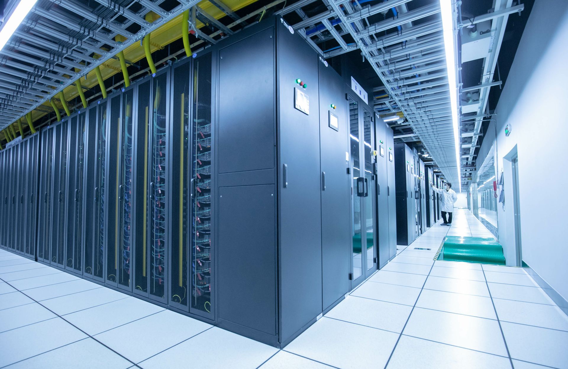 Rows of dark server racks in a data center; person in white coat visible in the distance.