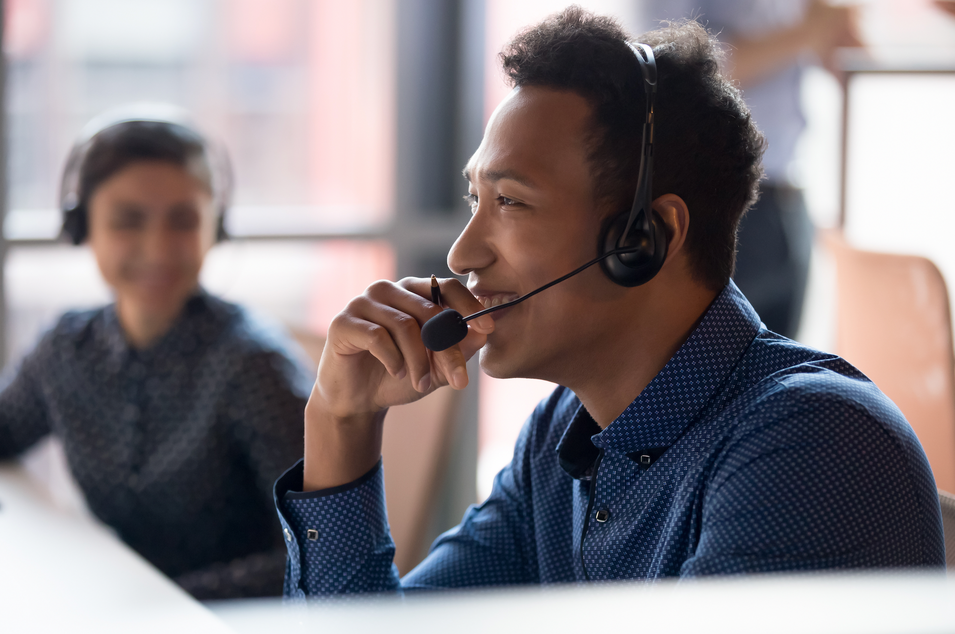 Man wearing headset smiles while speaking. Other person out of focus in background.