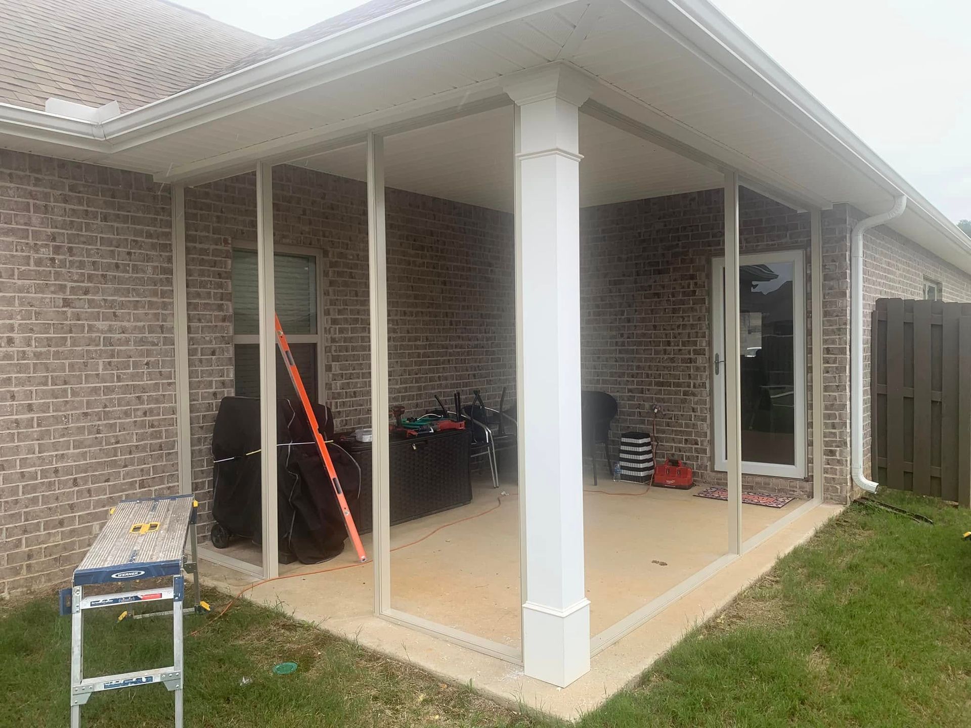 A patio area with a white column and metal framing being installed, a ladder, and brick walls of a residential home.