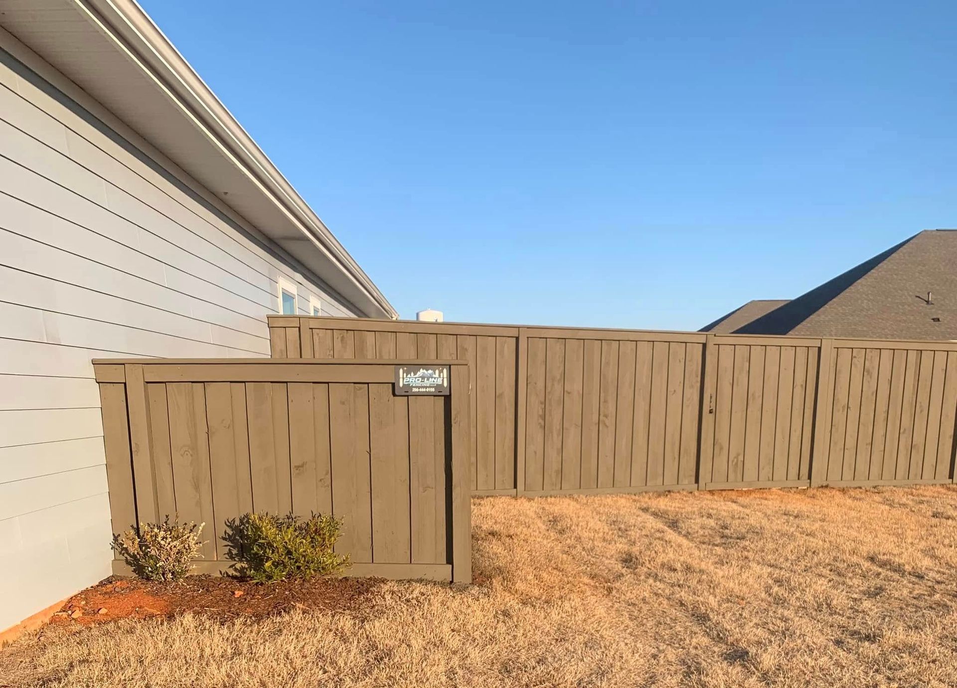 A beige wooden privacy fence runs alongside a house exterior with light gray horizontal siding against a blue sky.
