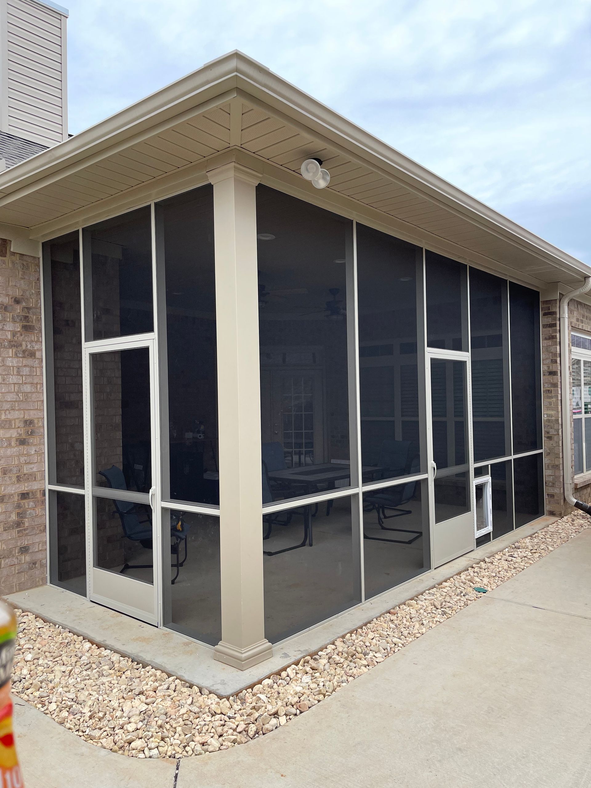 A screened-in patio with brick walls, a ceiling fan, and furniture visible inside, framed by a white metal screen system.