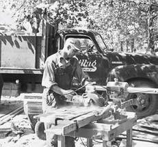 Vintage shot of Kreiling roofer masterfully cutting lumber with a radial arm saw, high lift truck in backdrop