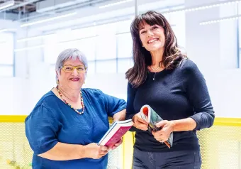 Duas mulheres estão de pé, uma ao lado da outra, segurando livros.