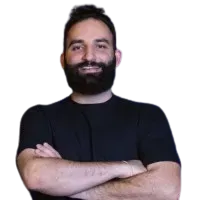 Um homem com barba está de pé com os braços cruzados e sorrindo.