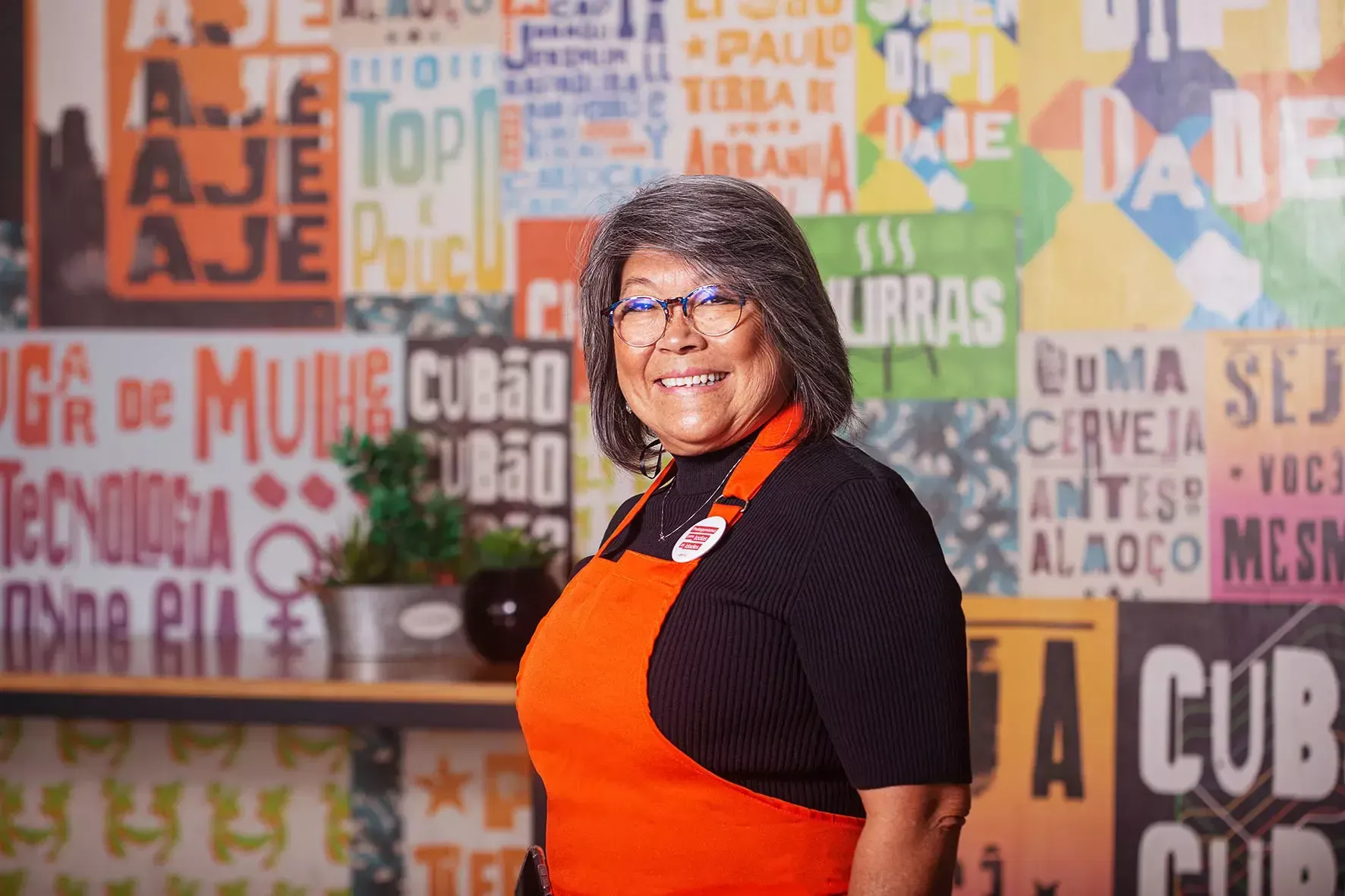 Uma mulher usando óculos e avental está parada em frente a uma parede colorida.