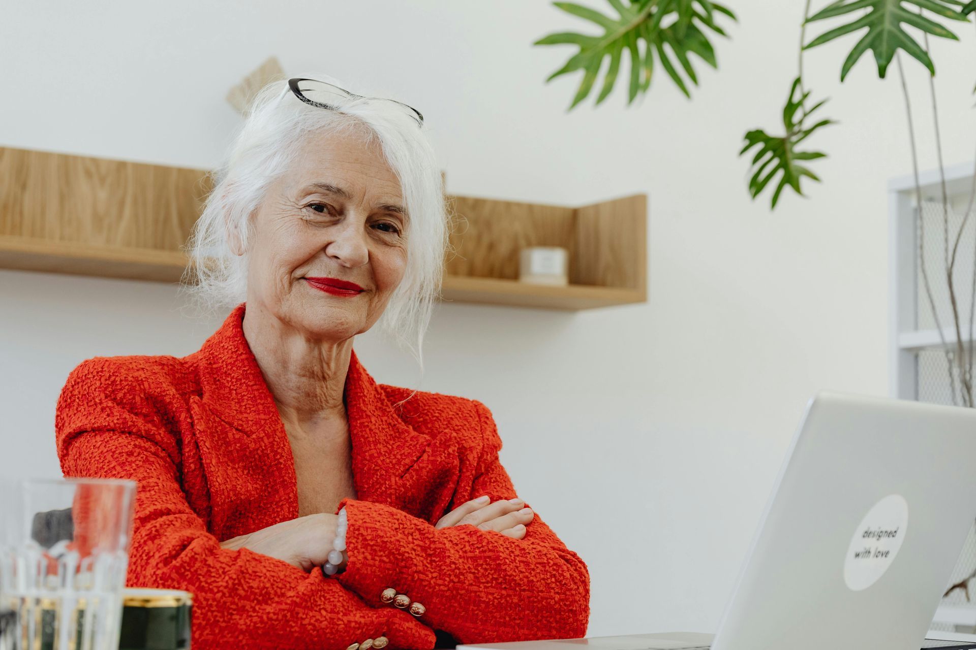Woman in red blazer, smiling, arms crossed, at a desk with a laptop and a plant in the background.