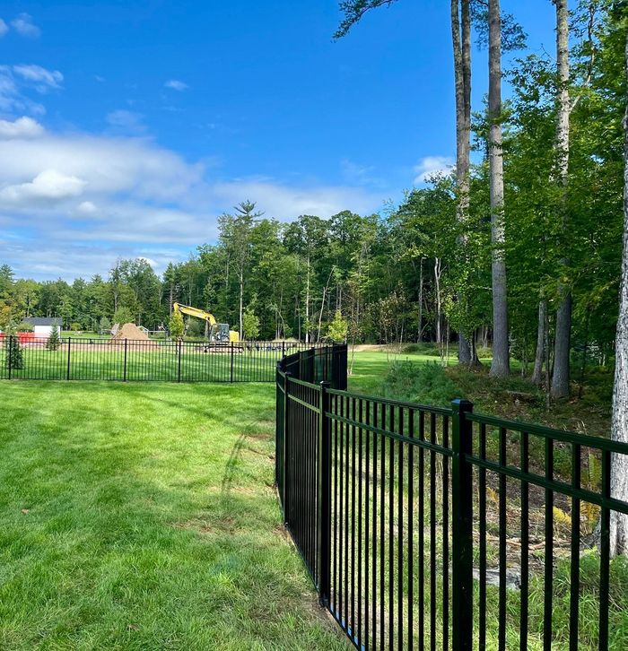 Two people constructing a wooden fence outdoors, one on a truck bed, in front of a wooded backdrop.
