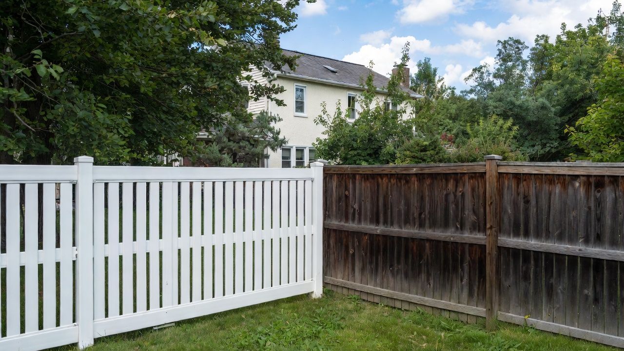 White picket fence next to a weathered brown wooden fence in a grassy yard, with a house and trees in the background.