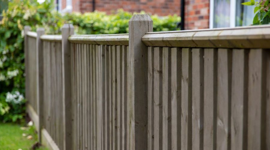 A wooden fence with vertical slats and square-topped posts stands in a garden near a house wall and greenery.