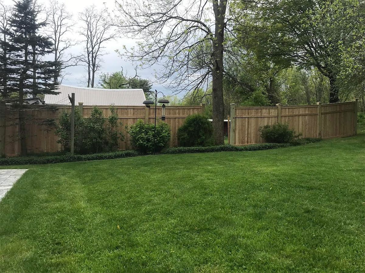 Backyard with a gray patio, white fence, lawn, and house under a blue sky with fluffy clouds.