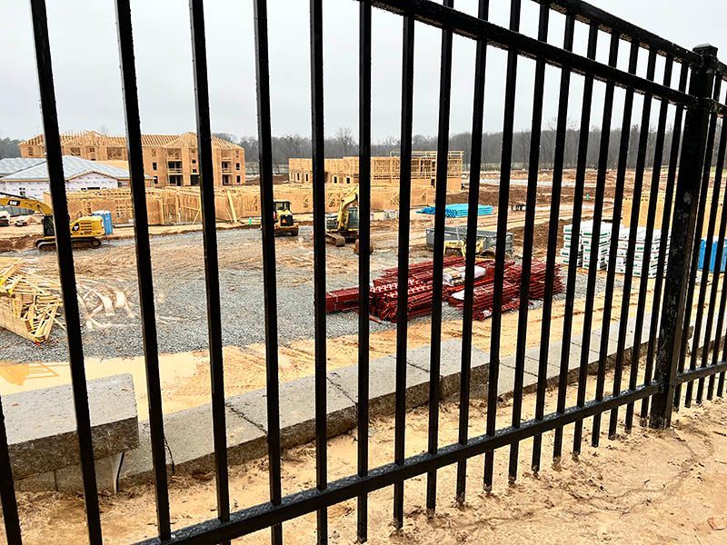 Construction site viewed through a black metal fence; heavy machinery, lumber, and unfinished buildings are visible.