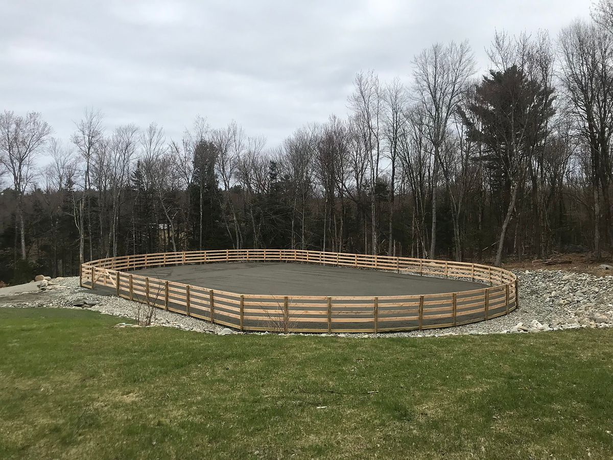 Wooden fence encloses a circular riding arena filled with sand, set in a grassy area near trees.