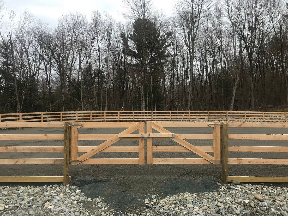 Wooden farm gate with diagonal bracing, open, leading to a gravel driveway and a treeline.