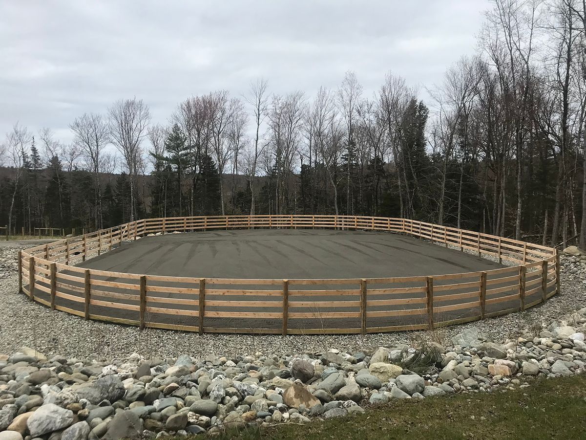 A circular horse riding arena with a wooden fence, dirt surface, and a rocky perimeter, set in a wooded area.
