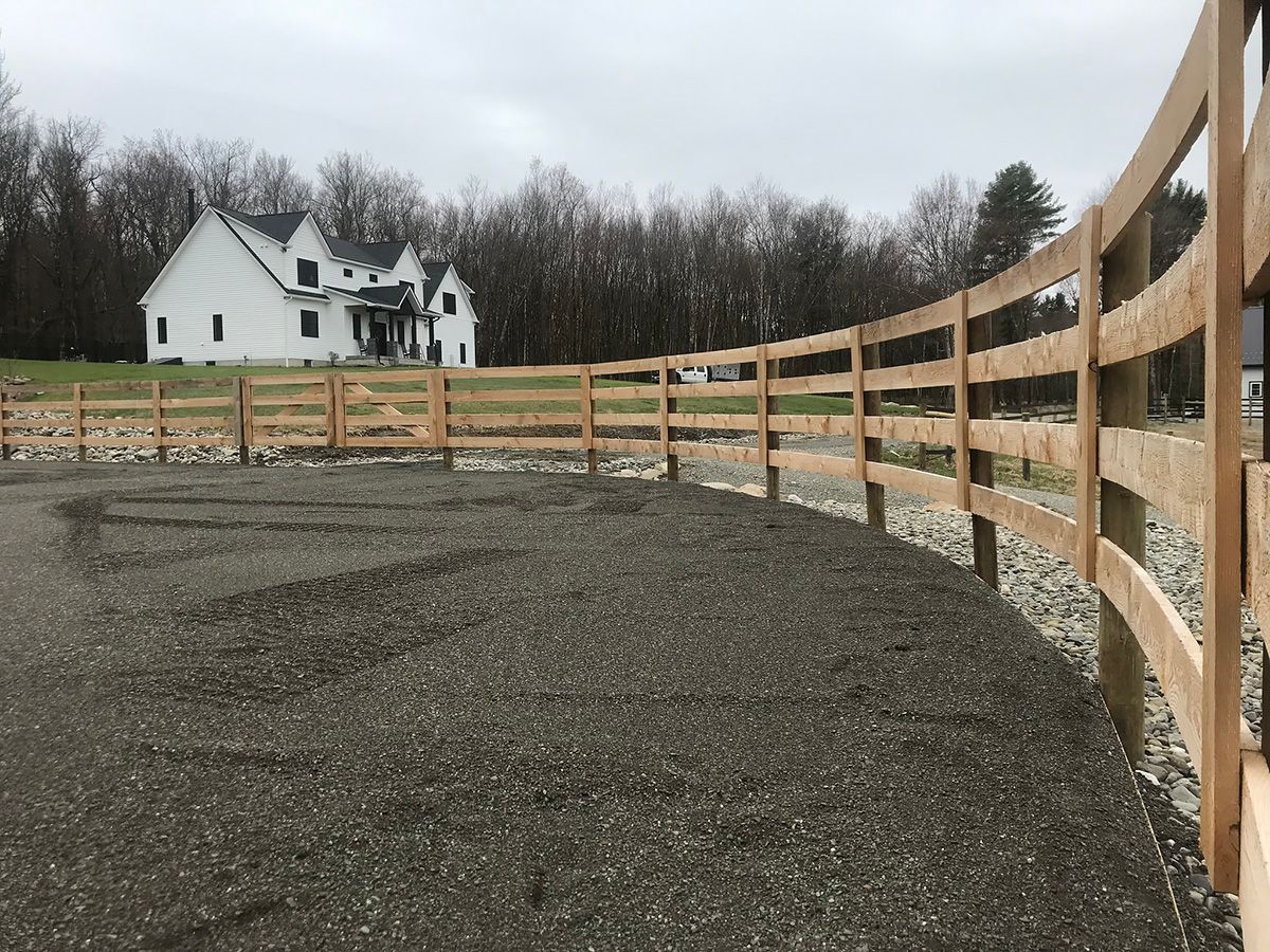 Wooden fence curves around gravel area in front of a white house on a cloudy day.