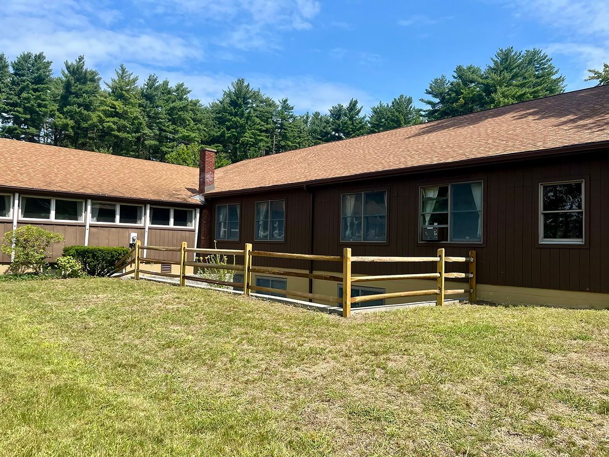 Brown building with a wooden fence in a grassy area, trees in the background, blue sky.