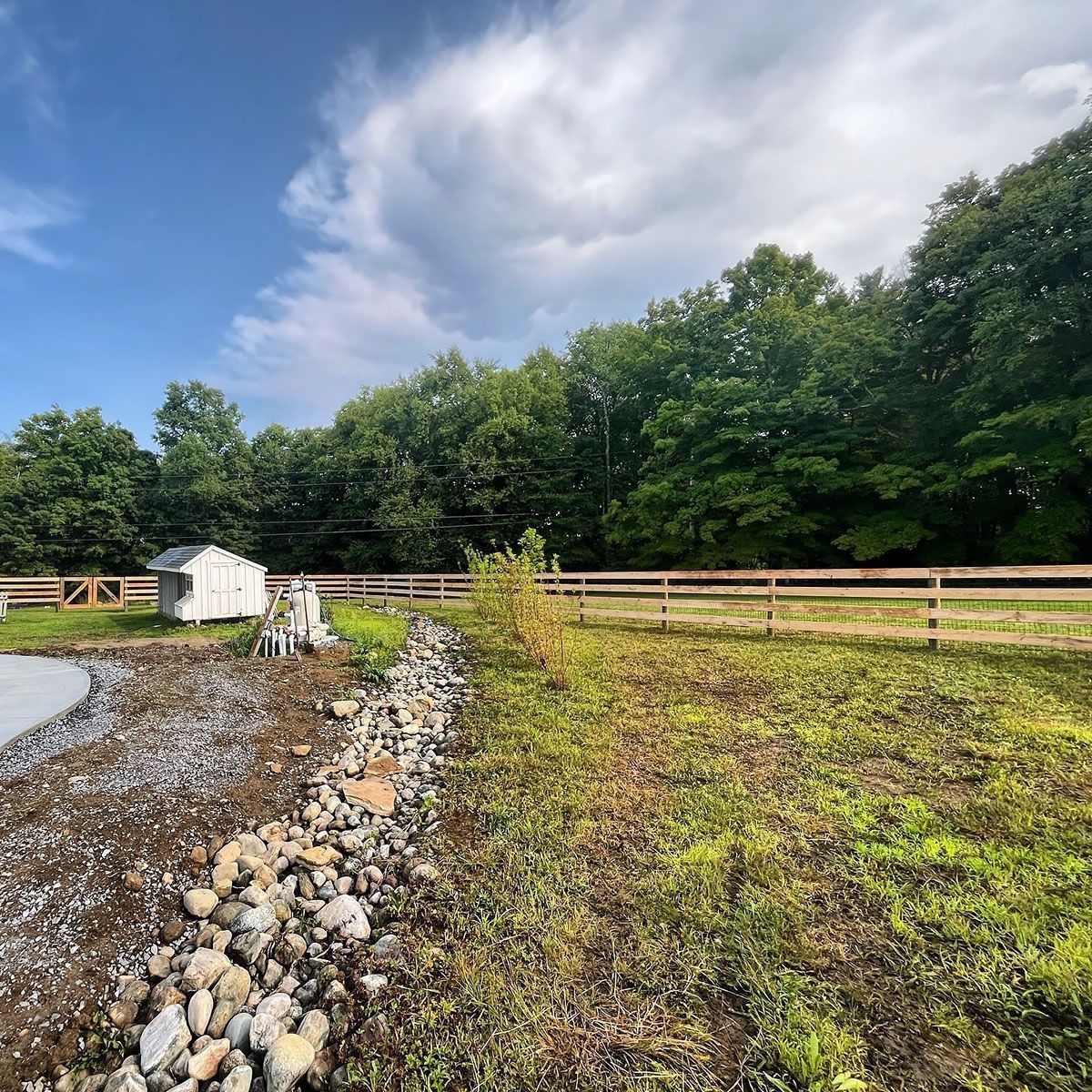 Lush green trees and a wooden fence border a grassy field, with a small shed and rocky shoreline. Cloudy sky.