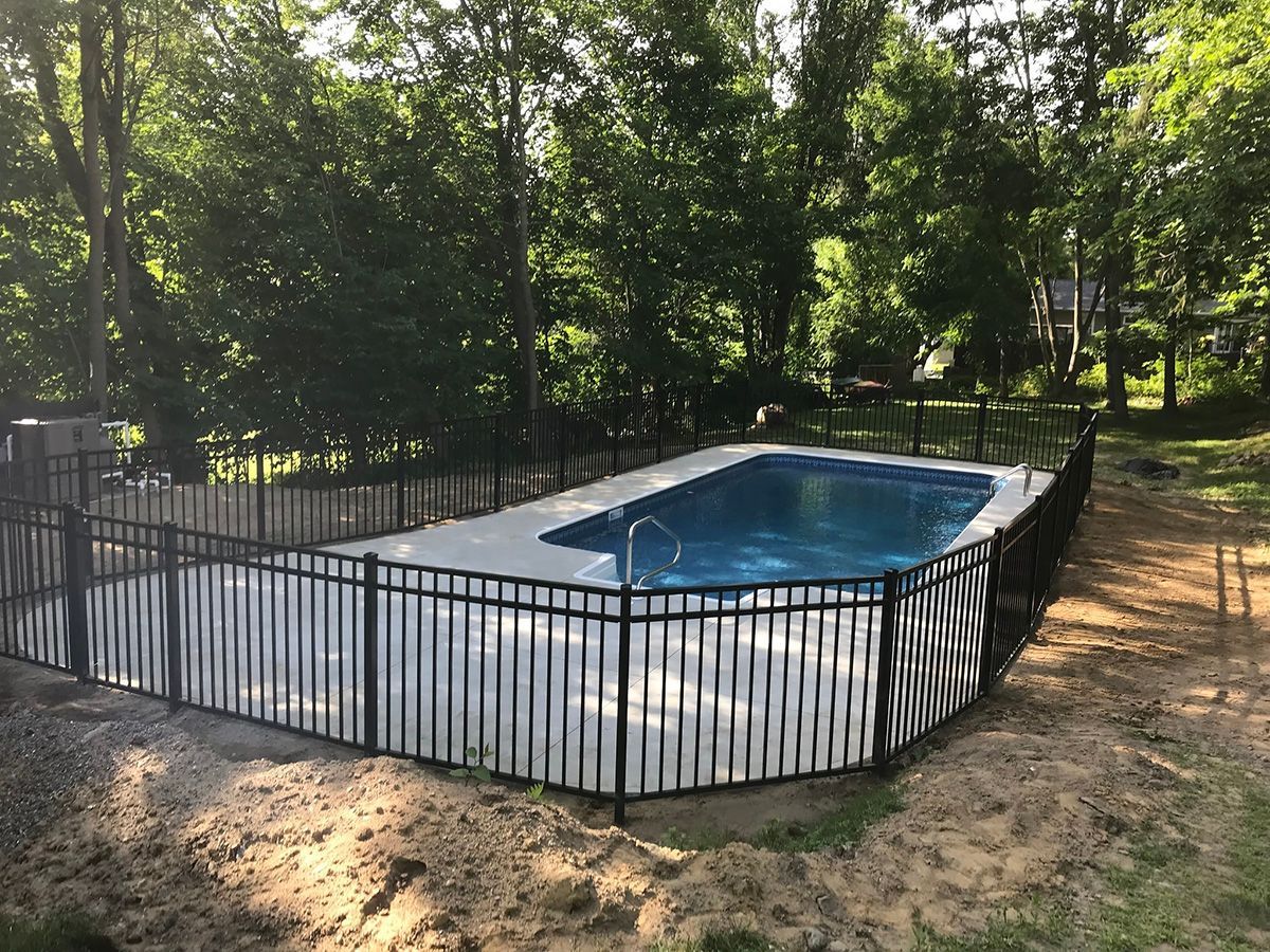 Fenced-in rectangular swimming pool. Black fence surrounds pool on sandy ground, with trees in the background.