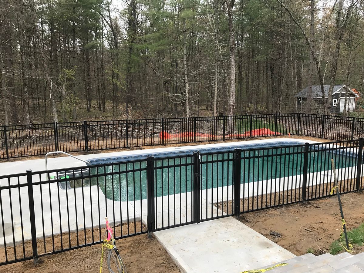 Black fenced swimming pool in wooded area. Concrete walkway leads to pool.