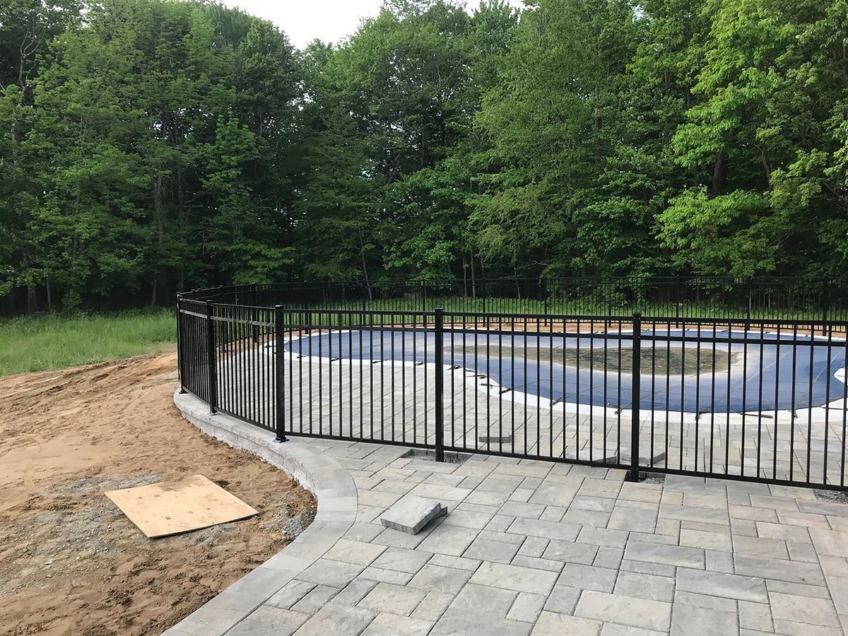 Black fence surrounds a pool covered with blue tarp, beside a stone patio and dirt, trees in the background.