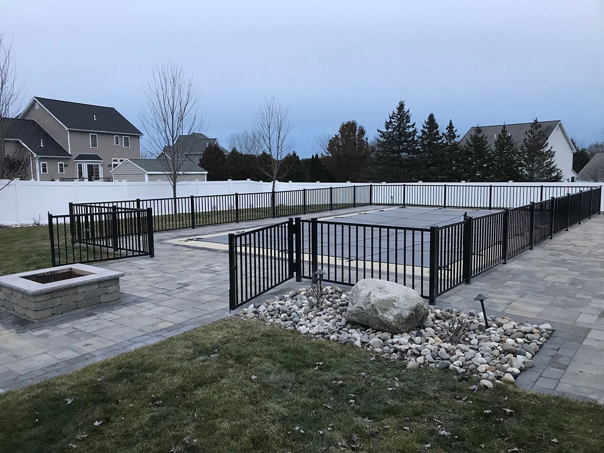 Black fence surrounds a pool on a stone patio with a fire pit, rock garden, and grassy lawn in a residential backyard.